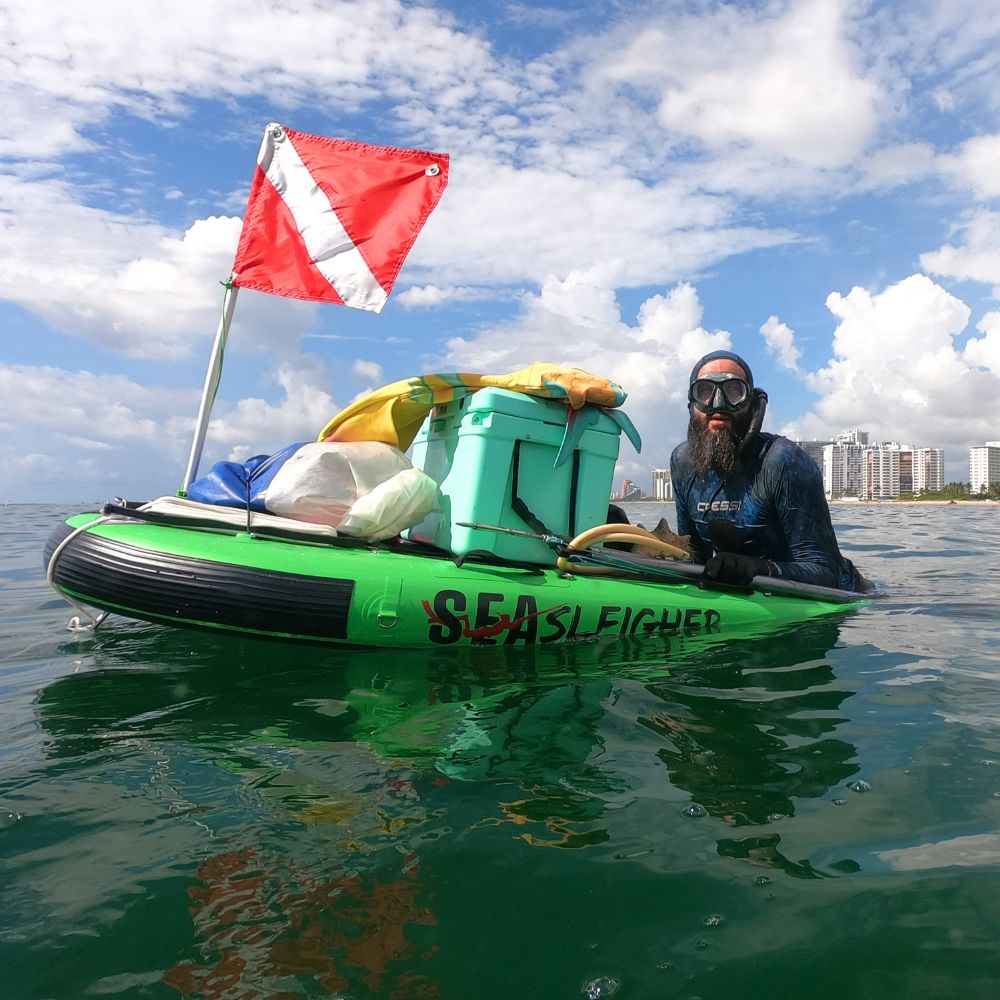 Squatch and Siren Sea Sleigher moray green inflatable dive raft on the water with a cooler, dive flag, other gear, and adult on top.